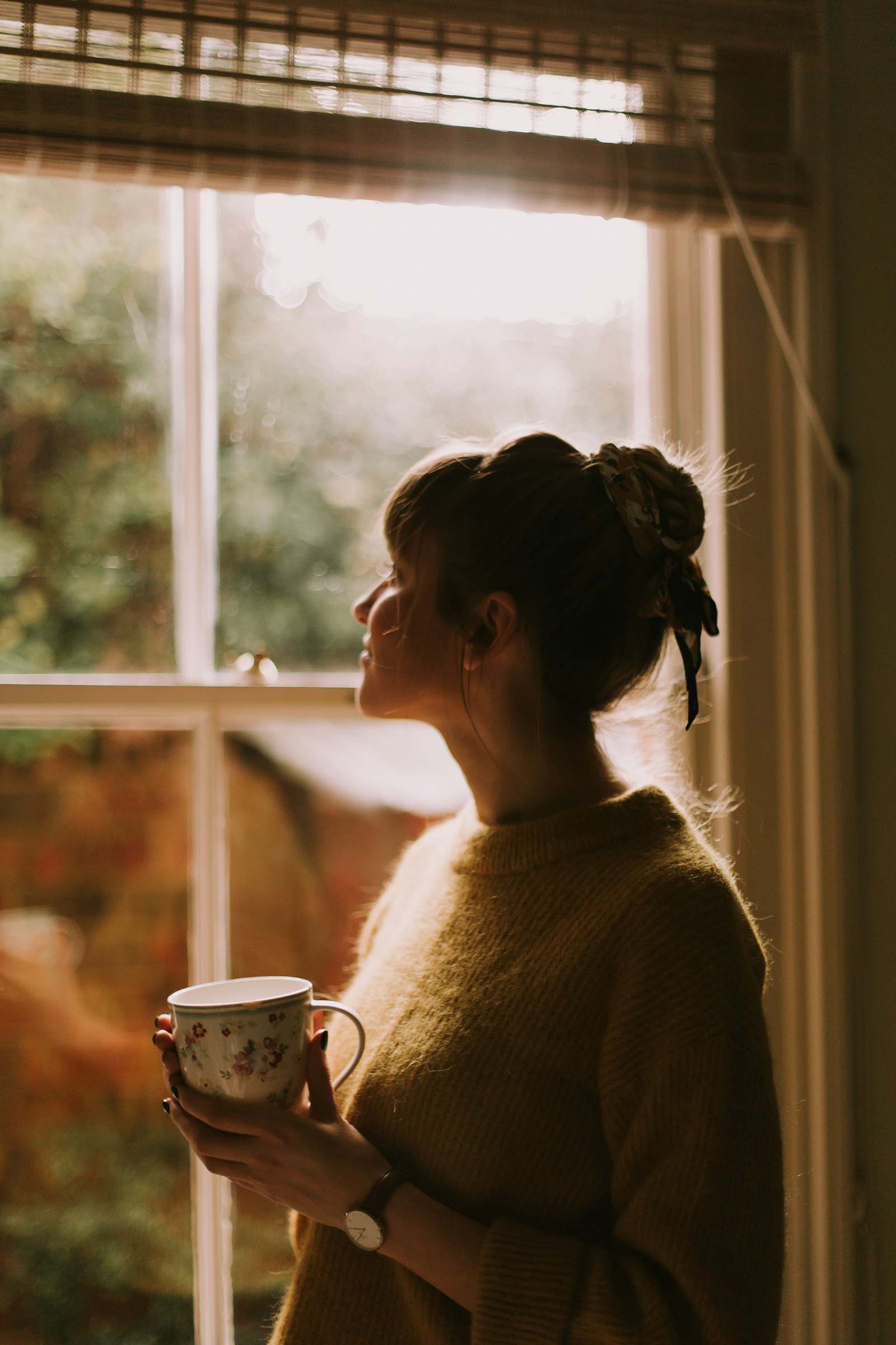 A woman alone by a window in golden light, holding a mug, looking out reflectively